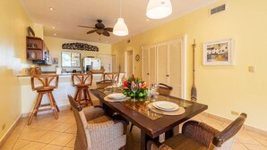 A dining area with a dark wooden table set for four, surrounded by wicker chairs. A ceiling fan and pendant lights are visible above the table. - Sunrise 7 - 2 Bedroom, Ocean View Condo (Tamarindo)
