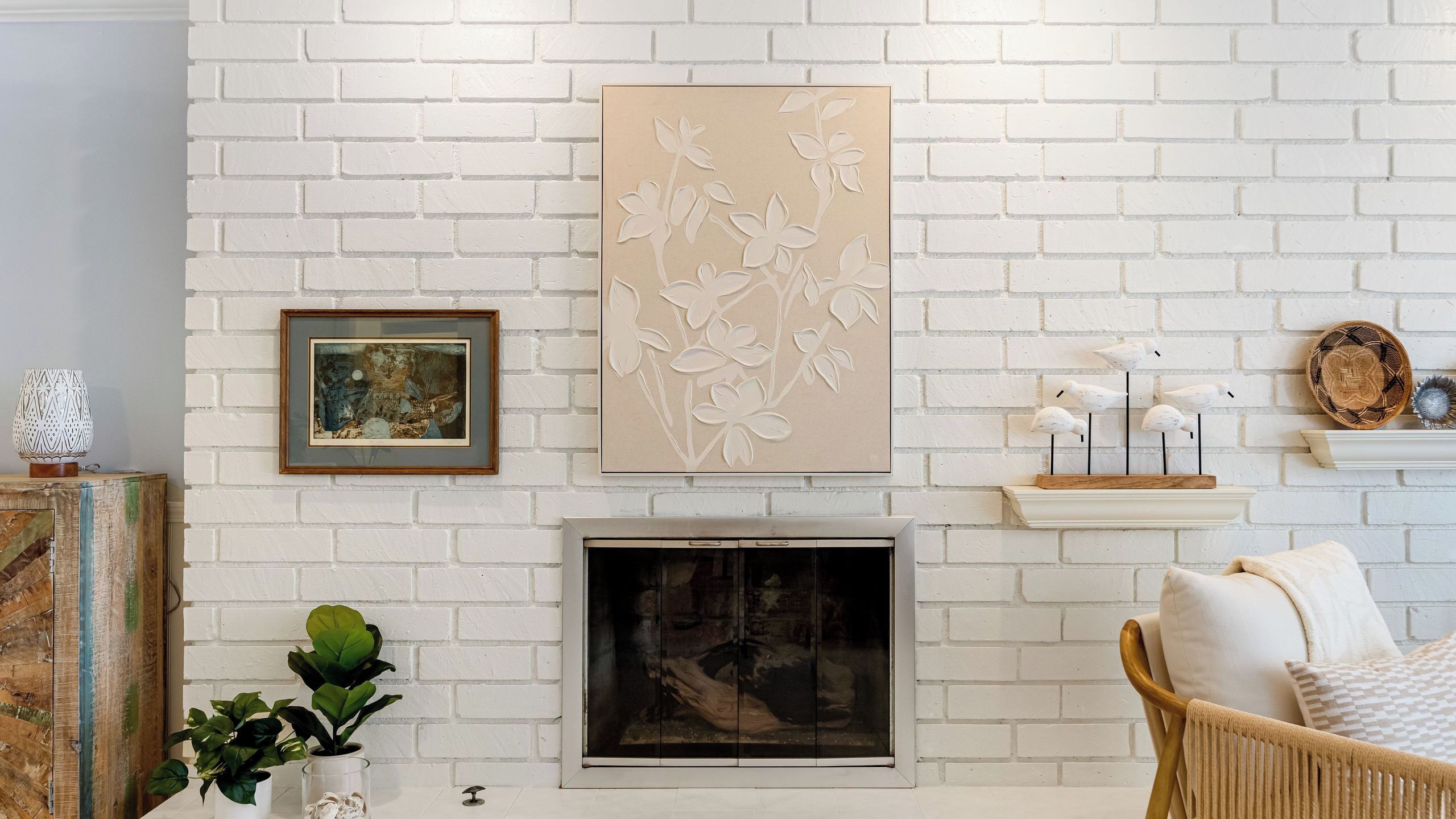Paredes de tijolos brancos com uma lareira, um quadro emoldurado, uma planta em vaso e uma cadeira de vime.