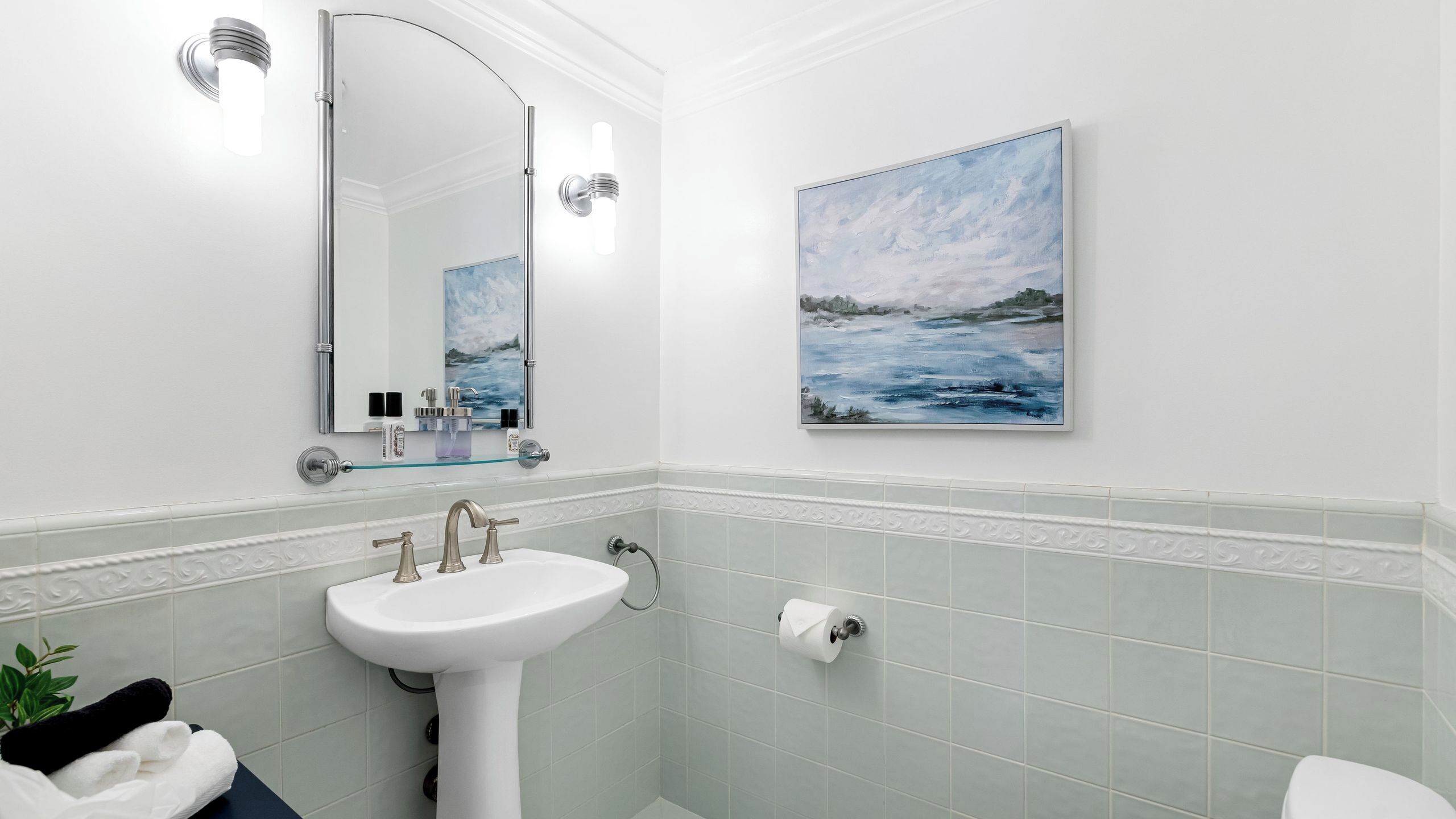 Ein Badezimmer mit einem weißen Waschbecken, einem Spiegel, einem Landschaftsbild und einer Toilette.