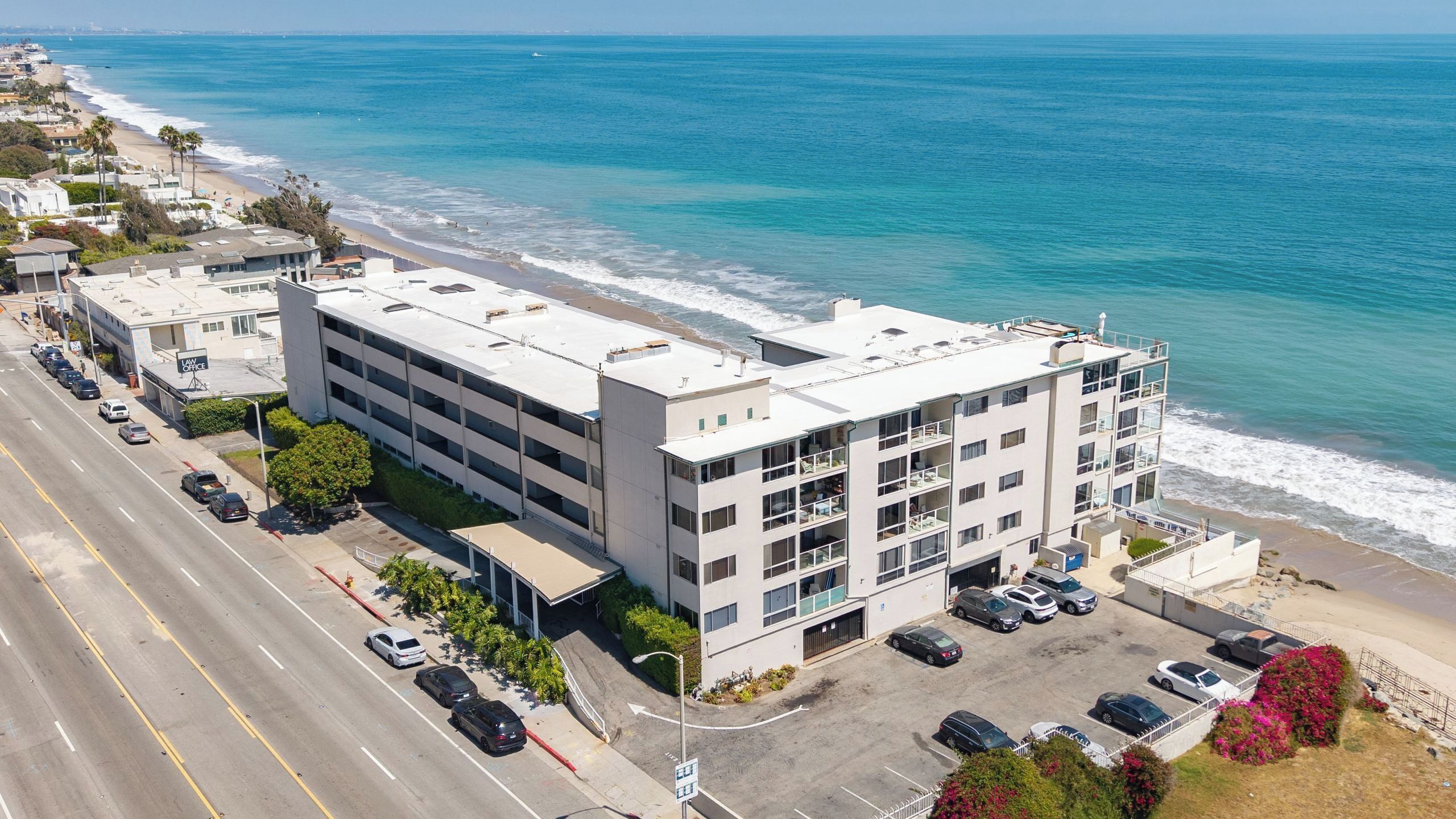 A multi-story building with a parking lot, adjacent to a beach and ocean.