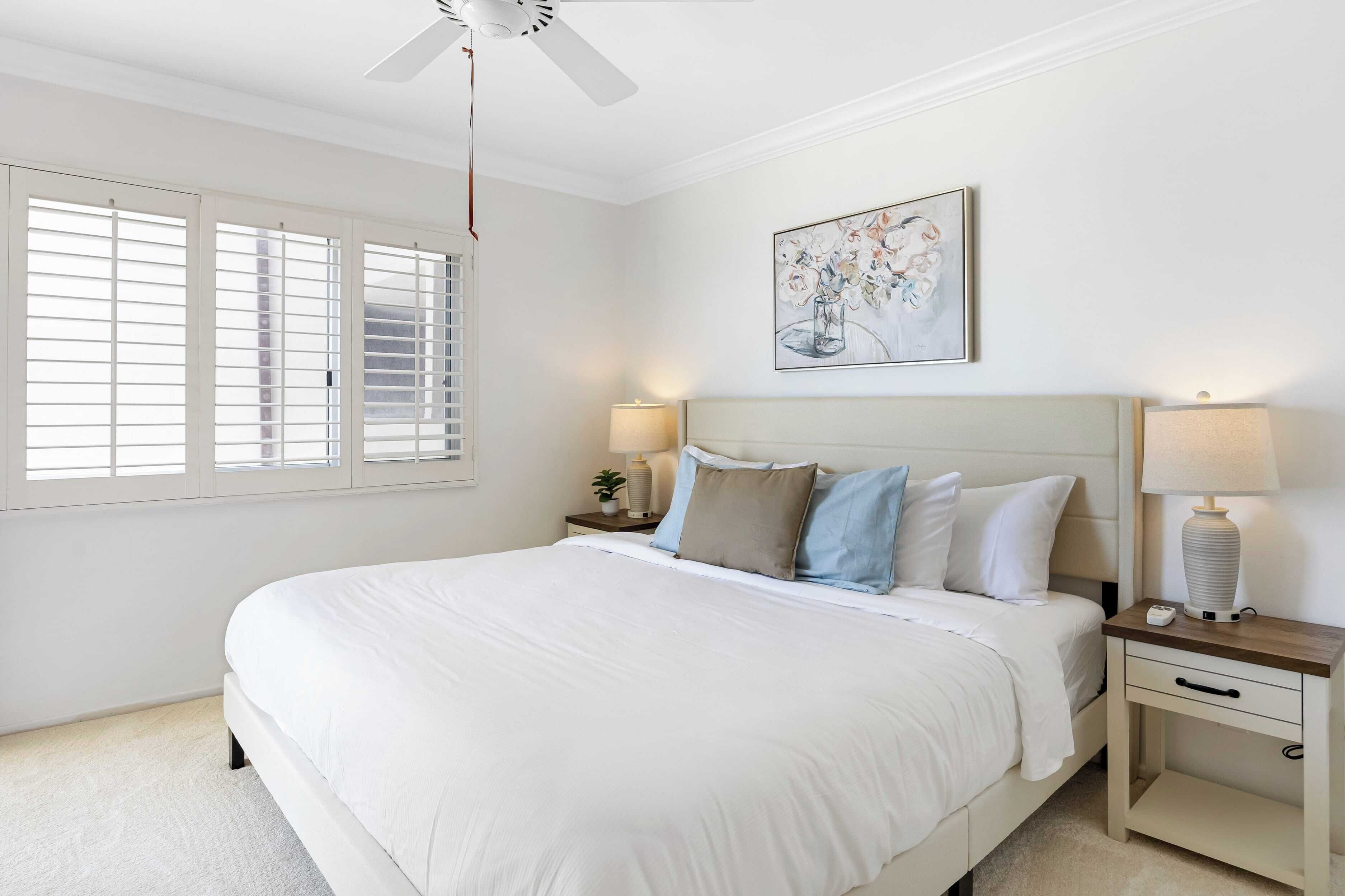 A bedroom with a large bed, two bedside tables with lamps, a ceiling fan, and a window with blinds.