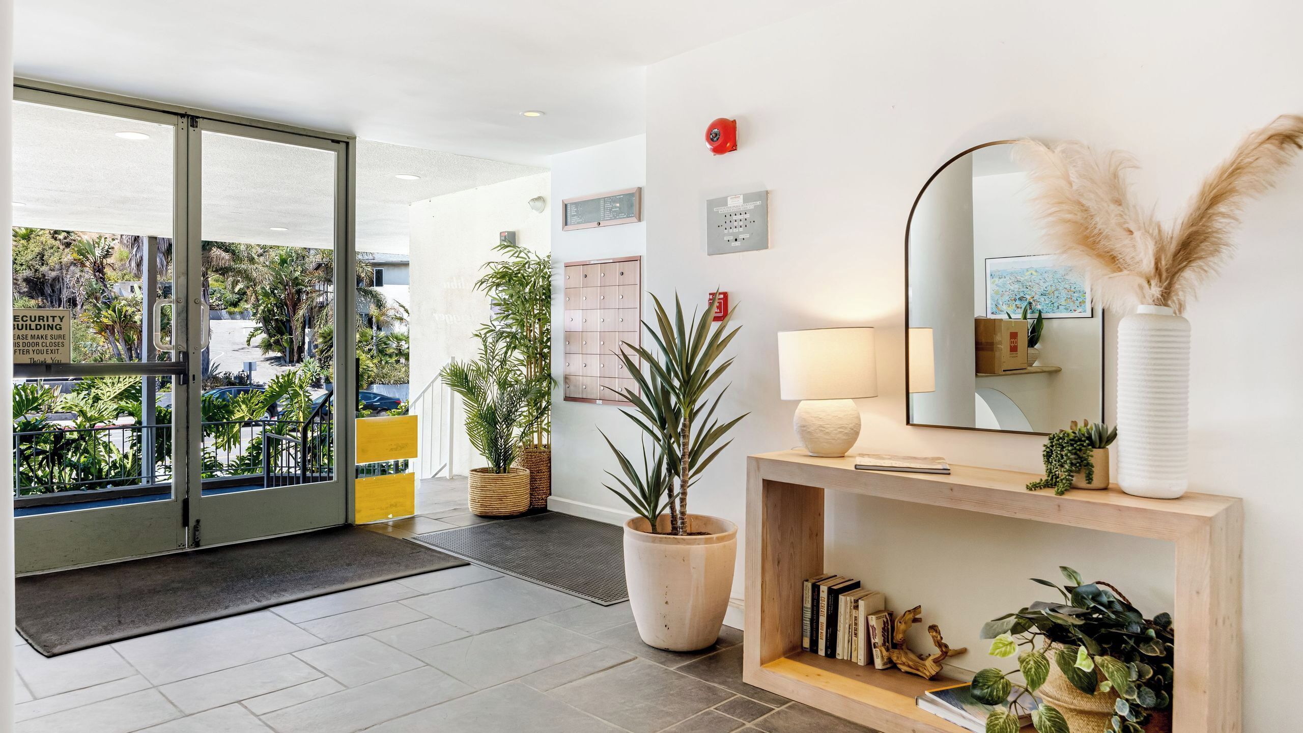 A hotel lobby with a large glass door, a reception desk, a mirror, a lamp, and a potted plant.