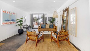 A modern living room with a sofa, two wicker chairs, a coffee table, and a large mirror. There is a poster on the wall and a potted plant. - Oceanfront Gem w/ Balcony, Pool, Hot Tub & Carbon Beach Access Bluewater Haven by Avantstay (Malibu)
