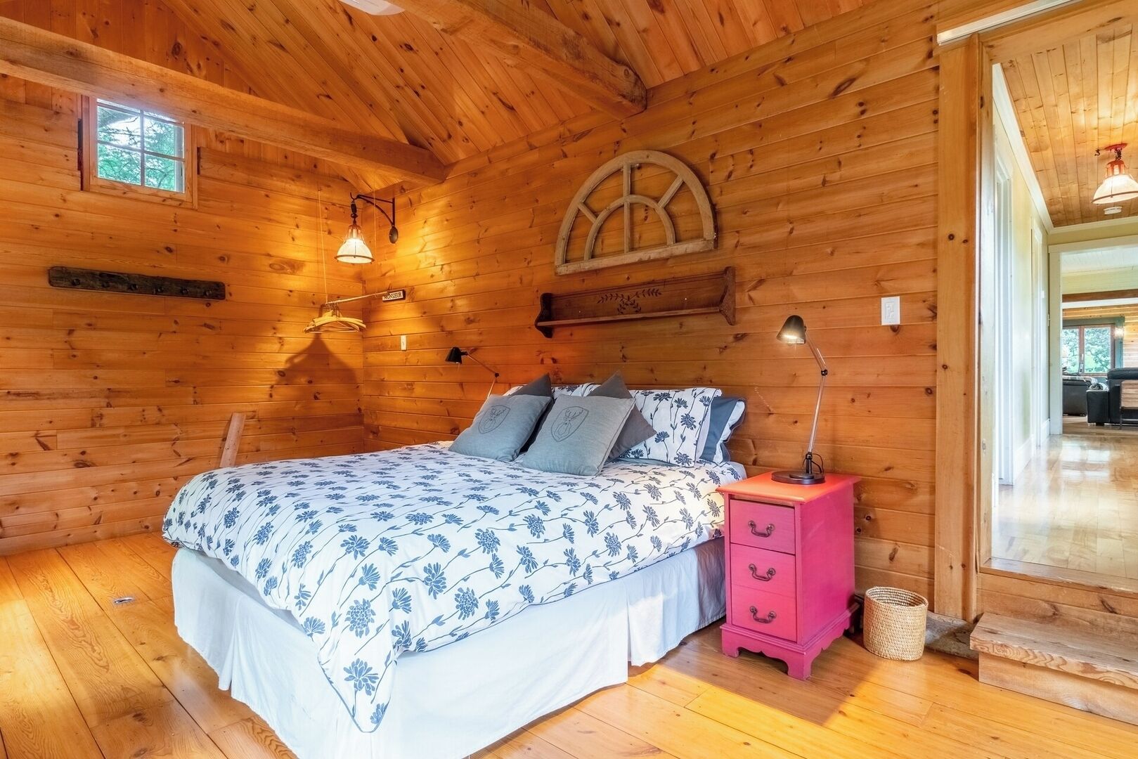A wooden cabin bedroom with a bed, bedside lamps, a pink nightstand, and a window.