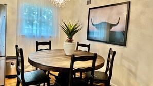 Un coin repas avec une table ronde en bois, quatre chaises, une plante en pot, un lustre et un tableau encadré au mur.