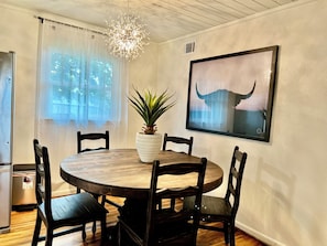 Een eethoek met een ronde houten tafel, vier stoelen, een potplant, een kroonluchter en een lijst met een afbeelding aan de muur.
