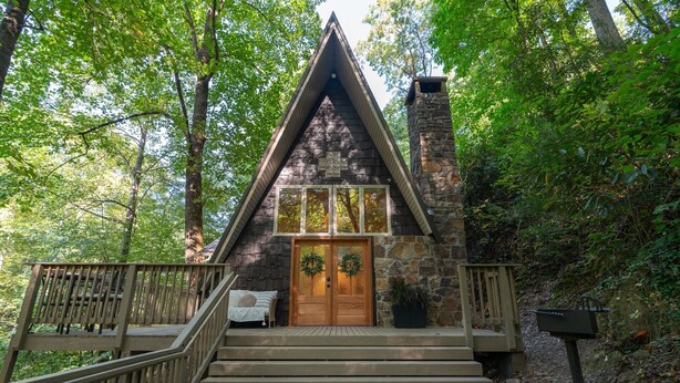 A cabin with a wooden front door, a stone chimney, and a wooden staircase leading up to the entrance. - Cozy A-Frame Retreat (Gatlinburg)