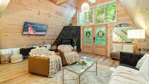 A cozy, wooden-floored living room with a fireplace, comfortable seating, and large windows offering a view of the outdoors. - Cozy A-Frame Retreat (Gatlinburg)
