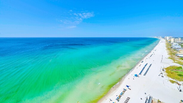 A beach with clear turquoise waters, a sandy shore, and buildings along the coastline.
