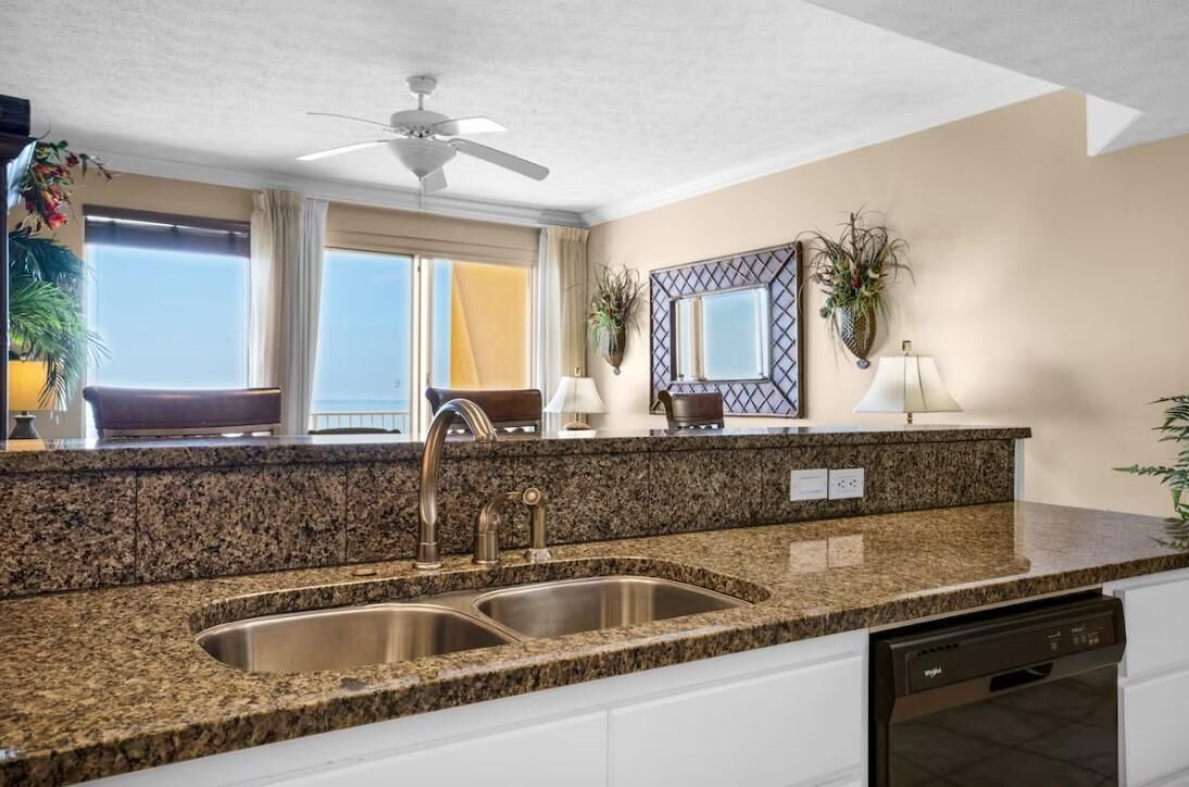 Una cocina moderna con encimeras de granito, lavamanos de acero inoxidable y lavavajillas. Hay un ventilador de techo, ventanas amplias con cortinas y un espejo decorativo sobre el lavamanos.