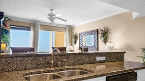 A modern kitchen with granite countertops, stainless steel sinks, and a dishwasher. There is a ceiling fan, large windows with curtains, and a decorative mirror above the sink.