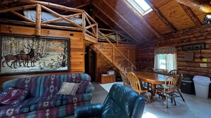 L’intérieur d’un chalet rustique, avec un escalier en bois, une table à manger entourée de chaises, un canapé et un tableau représentant des cerfs.