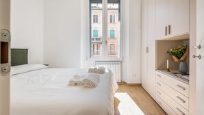 A bedroom with a bed, a window with a view of buildings, a wardrobe, and a bedside table with a vase and candle. - New NoLo - La Vista Apartments (Milan)