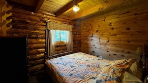 A log cabin bedroom with a bed, a window with curtains, and a wooden ceiling.