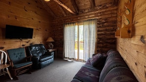 A log cabin interior with a wooden ceiling, a flat-screen TV mounted on the wall, a sofa, two chairs, and a table with a lamp.