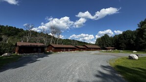 Filera de cabanes de fusta amb un camí de grava que condueix a un edifici de fusta, envoltat d'arbres i un cel serè.