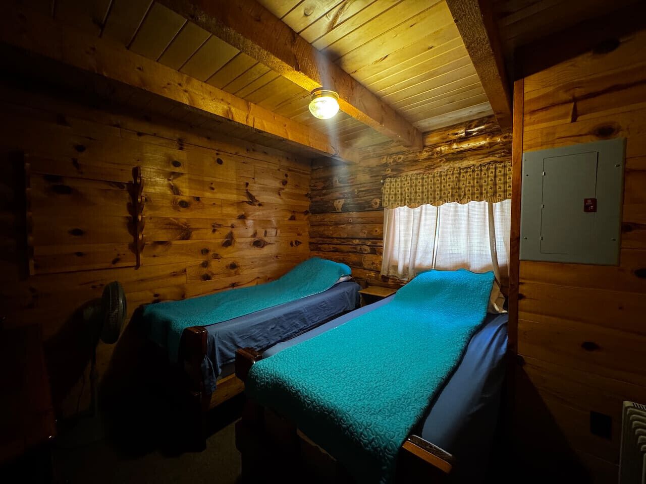 A cabin-style room with two beds, wooden walls, a ceiling light, and a power outlet.