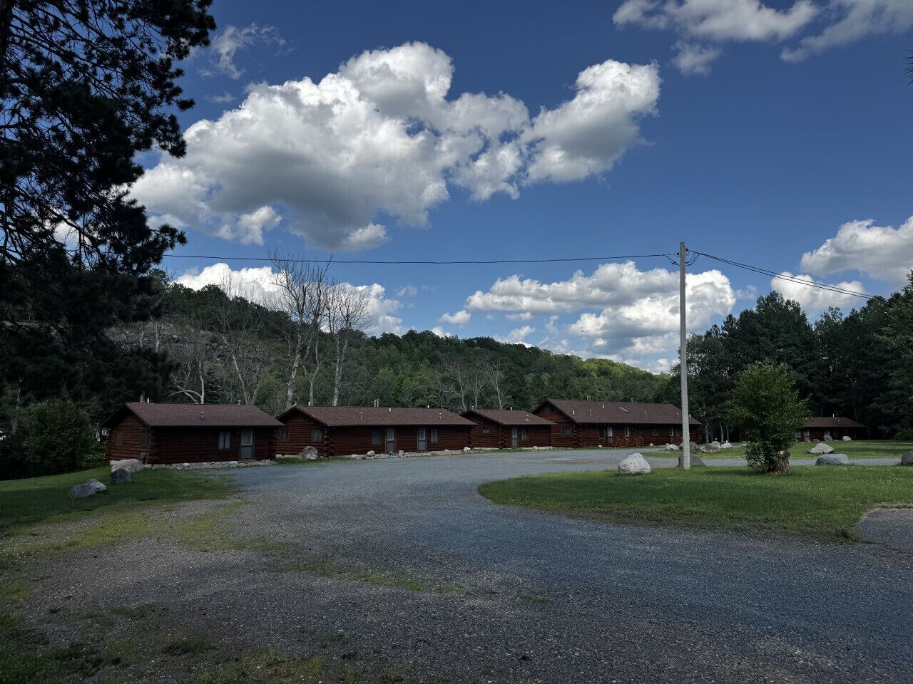 A row of log cabins with a gravel driveway leading to a parking area, surrounded by trees and a mountain backdrop.