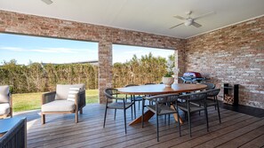 A covered patio with a brick wall, a round wooden table, wicker chairs, a grill, and a ceiling fan. - Holiday Crescent – Spacious, stylish retreat with pool, perfect (Berry)