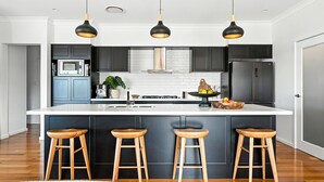 A modern kitchen with dark cabinetry, a white island with bar stools, and pendant lighting. - Holiday Crescent – Spacious, stylish retreat with pool, perfect (Berry)