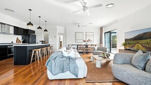 A modern kitchen with dark cabinets, a central island with stools, a dining area with a table and chairs, a living room with a sofa and a coffee table, and a large window with a vineyard view. - Holiday Crescent – Spacious, stylish retreat with pool, perfect (Berry)