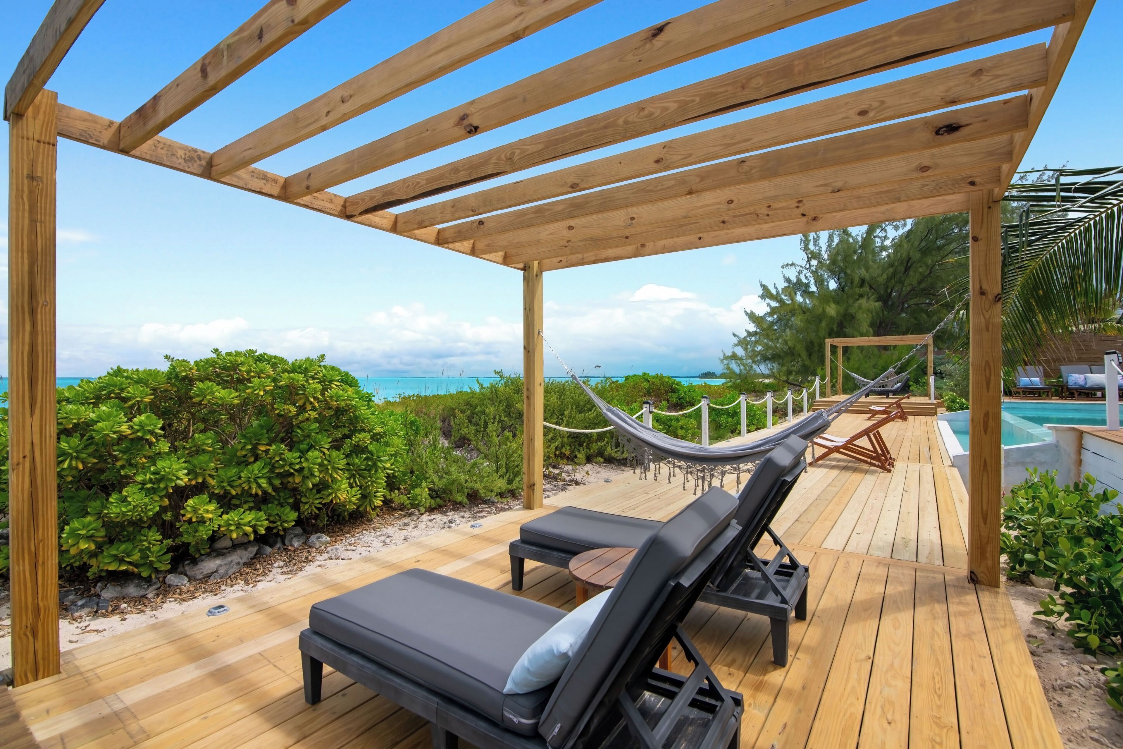 Een houten terras met lounge stoelen en een hangmat, met uitzicht op een zwembad en de oceaan.