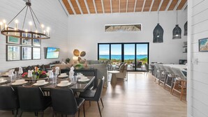 Un comedor amplio con una mesa larga dispuesta para una comida, un ventanal con vista al océano y una cocina moderna al fondo.