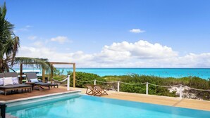 Zona de piscina con sillones de descanso y terraza de madera con vista a una playa y al océano.