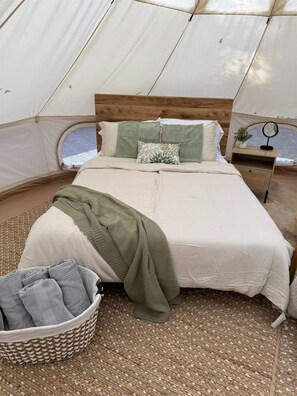 Una carpa con una cama, almohadas, una manta, una mesita con un espejo y una cesta de toallas.