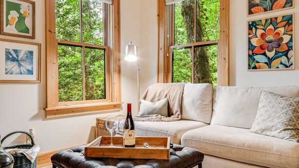 Un salon chaleureux avec un canapé, une table basse en bois sur laquelle sont posés une bouteille de vin et des verres, et de grandes fenêtres donnant sur un paysage verdoyant.