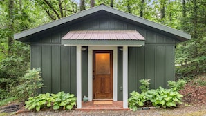 Un petit abri de jardin en bois, peint en vert, avec une porte marron et des colonnes blanches, entouré de verdure.