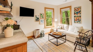 Un salon avec un canapé blanc, une table basse et un téléviseur fixé au mur. Il y a deux fenêtres avec des cadres en bois, donnant sur un paysage verdoyant.