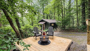 Une terrasse avec un foyer, deux fauteuils Adirondack et une petite structure en bois, le tout situé dans une zone boisée.