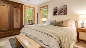 Une chambre avec une grande armoire en bois, un lit recouvert d’une couvre-lit à motifs, deux fenêtres avec des stores, un tableau accroché au mur et une table de chevet avec une lampe.