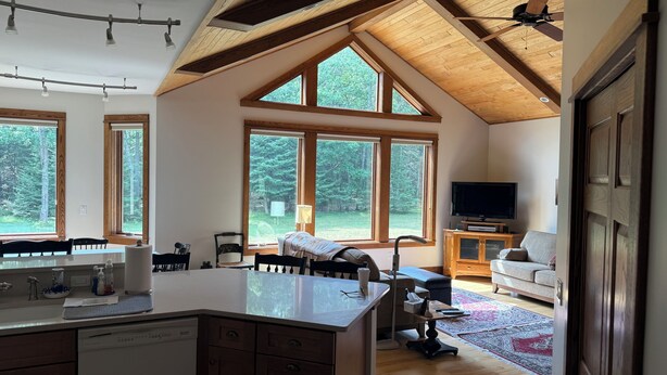 A spacious kitchen with a large island, wooden ceiling beams, and large windows offering a view of the forest.
