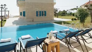 Un espace piscine avec des chaises longues et une table, un grand bâtiment aux volets bleus et un ciel dégagé.