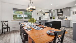 A modern kitchen with a central island, a dining area with a wooden table, and a view of the outdoors through a large window.