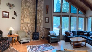 A cozy living room with a stone wall, a fireplace, and large windows offering a view of trees. - Black Butte Ranch `Trip House` On the Golf Course (Sisters)
