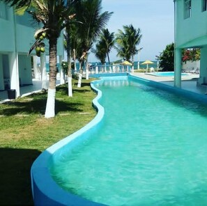 En swimmingpool med buet kant, omgivet af palmer og en bygning med hvid facade.