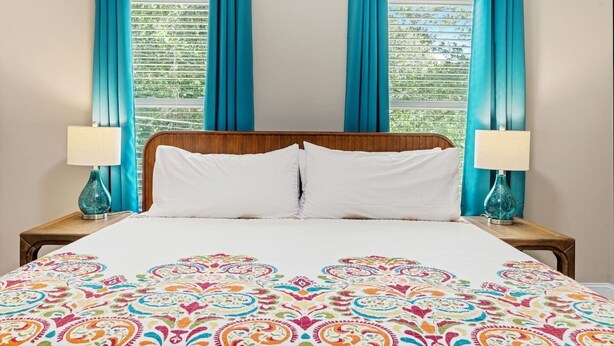 A hotel room with a wooden headboard, white bedding, and a colorful patterned bedspread. There are two bedside tables with lamps and a window with blue curtains.