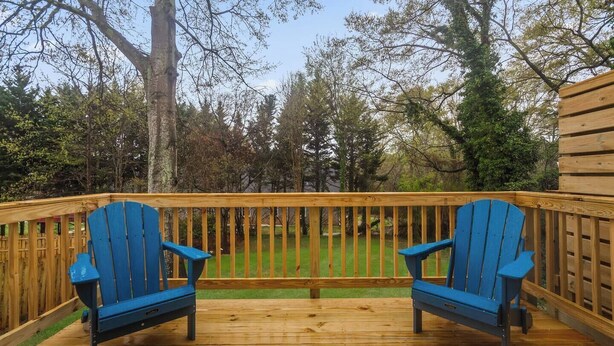 Una terraza de madera con dos sillas Adirondack azules enfrentadas, rodeada por un barandal de madera y un jardín con césped y árboles al fondo.