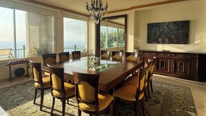 Une salle à manger spacieuse, avec une grande table en bois et des chaises recouvertes d’un tissu jaune, un lustre et une vue sur l’extérieur.