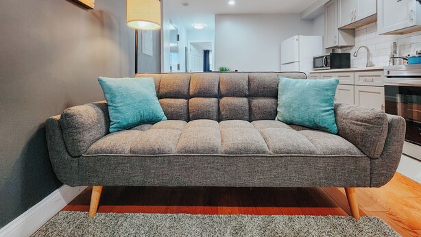 A modern living room with a grey sofa, teal cushions, a floor lamp, and a kitchen in the background. - 2 King Beds / 55" TV / Rainfall Shower / W/D (Philadelphia)