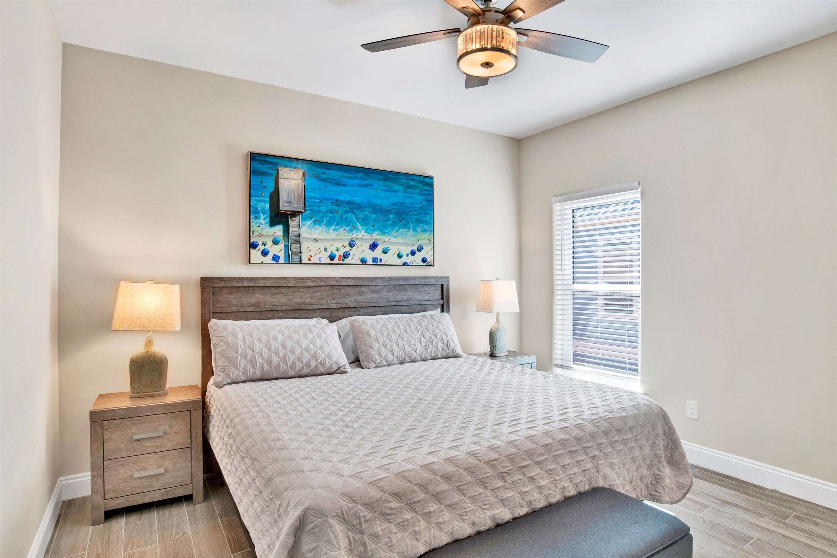 A bedroom with a bed, nightstands, a ceiling fan, a painting, and a window with blinds.