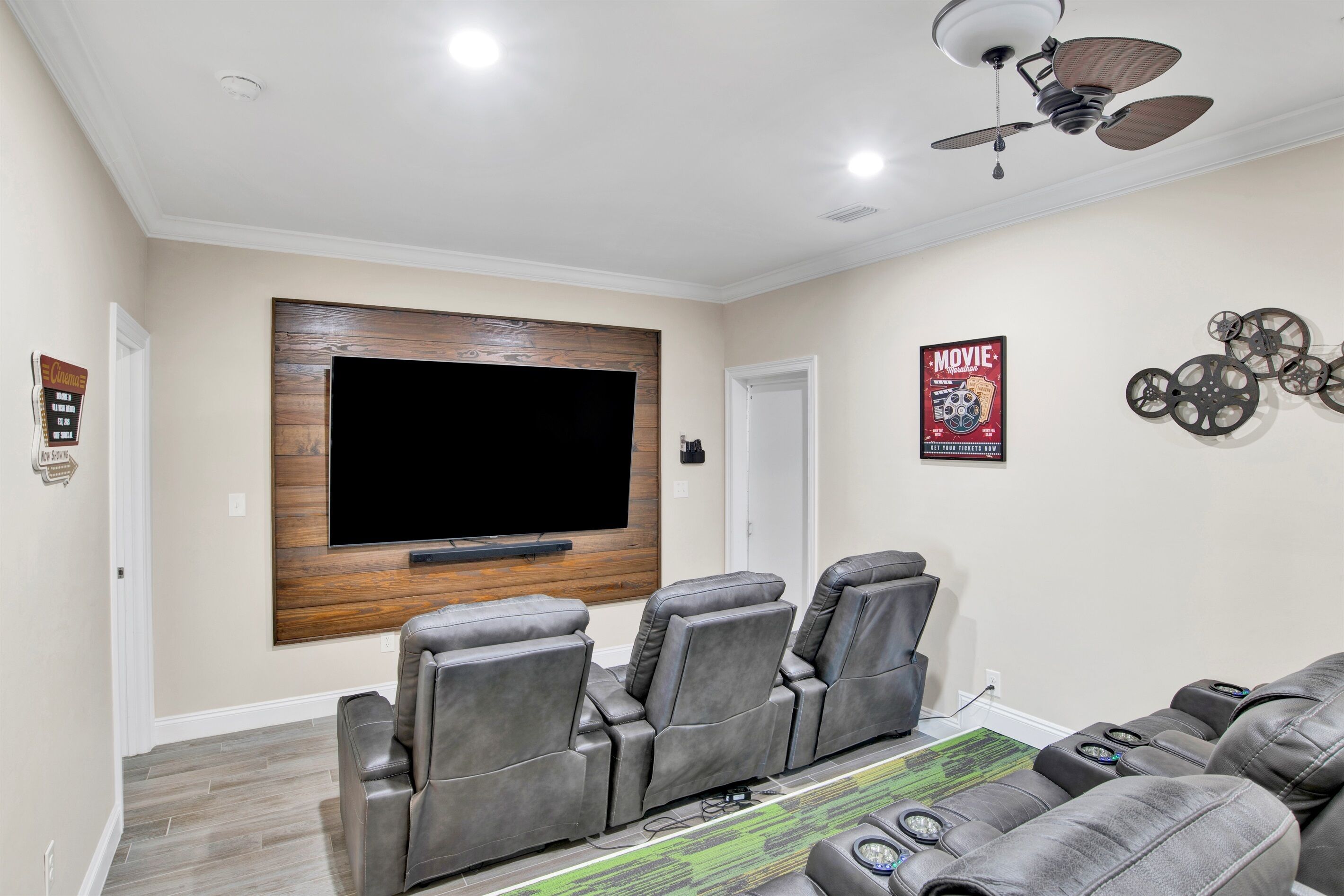 A home theater room with a large flat-screen TV mounted on a wooden panel, gray reclining chairs, a ceiling fan, and a poster on the wall.