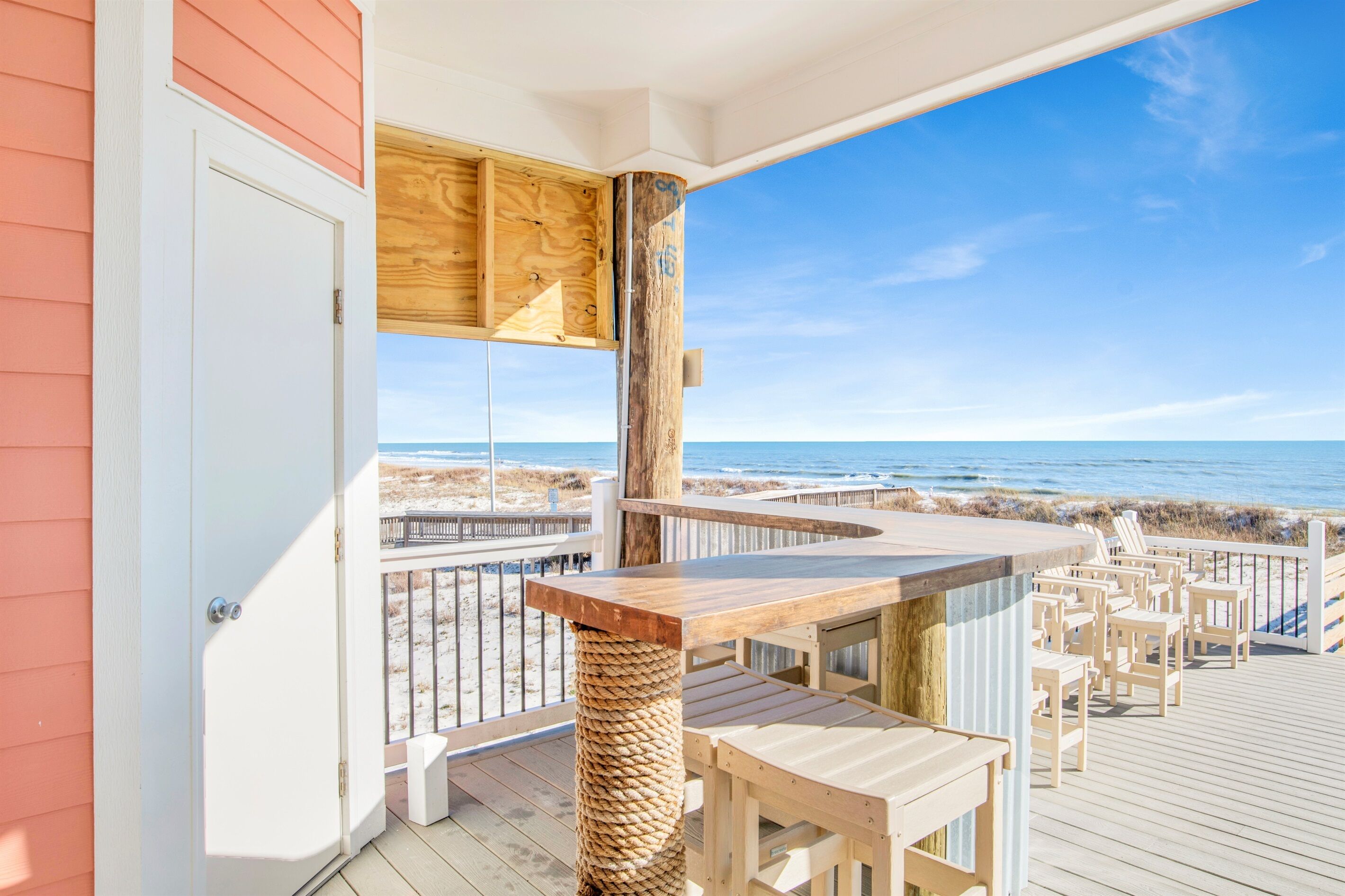 Terraza de madera con zona de bar y asientos, con vista a una playa.