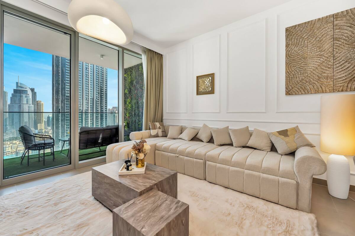 A modern living room with a large sofa, a coffee table, and a view of the cityscape through floor-to-ceiling windows.