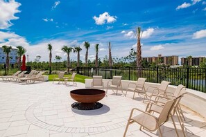 An outdoor lounge area with lounge chairs and a fire pit, surrounded by palm trees and a fence.