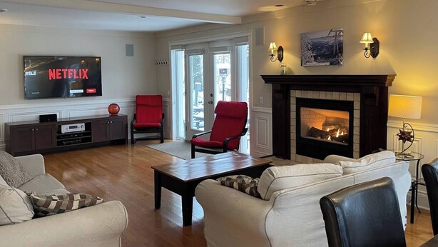 A living room with a fireplace, a television displaying 'NETFLIX', and a door leading outside. - Direct Bike Access, WaterPark, S’mores nights (Bromont)