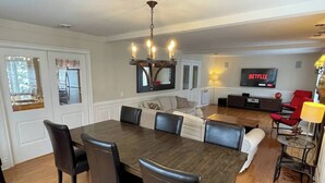 A dining area with a wooden table and black chairs, a living room with a sofa and a TV displaying 'NETFLIX', and a kitchen visible through a window. - Direct Bike Access, WaterPark, S’mores nights (Bromont)